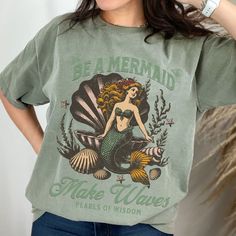 Dive into summer style with our Vintage Mermaid shirt! Featuring a stunning mermaid perched on a clam shell, surrounded by whimsical starfish, shells, and seaweed, this tee exudes retro seaside charm. With empowering messages like "Be a Mermaid, Make Waves" and "Pearls of Wisdom," it's more than just a shirt--it's a source of motivation and positivity. Whether you're soaking up the sun on a beach vacation or simply craving some summer vibes all year round, this sea siren shirt is the perfect cho Green Crew Neck Beach Shirt, Cotton Mermaid Top For Summer, Green Graphic Tee For Beach, Green Graphic Tee For The Beach, Green Screen Printed Tops For Beach Season, Green Graphic Tee For Beach Season, Green T-shirt With Screen Print For Beach Season, Green Screen Print Tops For Beach Season, Green Screen Print T-shirt For Beach Season