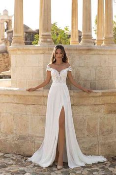 a woman standing in front of a stone wall wearing a white dress with an off the shoulder slit