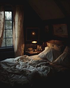 an unmade bed in a dark room with curtains on the window sill and a lamp