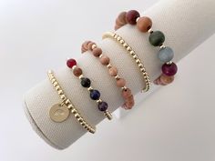 four bracelets with different colored stones and gold charms on each one arm, sitting on top of a white cloth