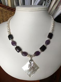 The focal point of this necklace is a rectangular pendant of white Mother of Pearls, which was handcrafted in Bali. The strand combines faceted ovals and faceted of rectangles of amethyst, grey freshwater pearls and silver accents. The necklace is 18 inches long, is strung on jeweler's wire and has a sterling silver lobster clasp. Item number 0519-SN8 White Amethyst Pendant Jewelry, Elegant Purple Rectangular Necklace, Elegant Rectangular Gemstone Beads Jewelry, Faceted Silver Pearl Necklace For Gift, Spiritual Rectangular Gemstone Beads Jewelry, White Amethyst Pendant Necklace, Elegant Purple Necklace, Handmade Purple Rectangular Necklace, Rectangular Purple Amethyst Necklaces