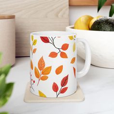 a white mug with orange and yellow leaves on it next to a bowl of fruit