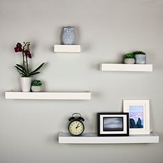 three white floating shelves with pictures, plants and an alarm clock on one shelf in front of the wall