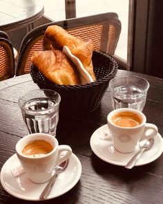 two cups of coffee sit on a table with pastries in the basket behind them