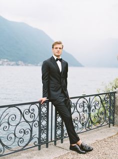 a man in a tuxedo leaning on a railing near the water and mountains