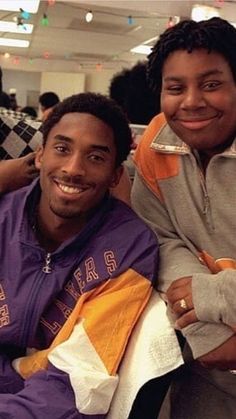 two young men sitting next to each other in a room with people standing around them