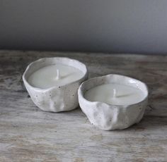 two white candles sitting on top of a wooden table next to each other, one with a lit candle in it