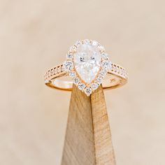 an engagement ring with a pear shaped diamond surrounded by small round diamonds on a wooden stand