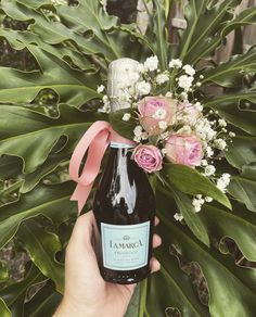 a person holding a bottle of wine in front of flowers and greenery with a pink ribbon