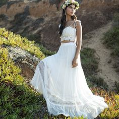 a woman standing on top of a lush green hillside wearing a white dress and flower crown