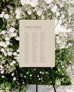 a sign that says take a seat in front of some bushes and white flowers on the ground