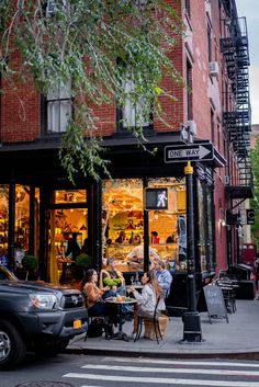 people sitting at tables in front of a store on a city street with cars parked nearby