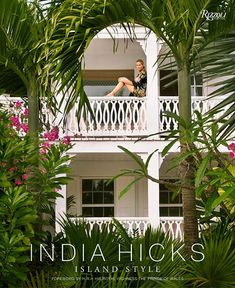 a woman standing on the balcony of a white house surrounded by palm trees and flowers