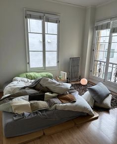 an unmade bed sitting on top of a hard wood floor next to two windows