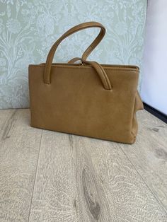 This is a stylish ladies handbag in a camel or beige leather-look material that has a smooth texture. It has a rectangular shape and a vintage look that evokes the elegance of the mid-century. The inside is lined with black satin that feels soft and luxurious. It has two side zipped pockets that are spacious and secure, and a middle section with a small zipped pocket that is perfect for storing small items. The frame, clasp and feet have gold accents that contrast beautifully with the neutral collar of the bag. The size is 28 cm wide, 16.5 cm high and 9 cm deep at the base, making it a versatile accessory that can fit many essentials.  The frame has the mark of Elbief England, a company that made frames for high quality British handbags from the 1940s to the 1970s. The mark is a sign of cr Beige Rectangular Satchel With Hasp Closure, Rectangular Beige Satchel With Hasp Closure, Vintage Brown Box Bag For Everyday, Chic Camel Satchel For Shopping, Vintage Square Box Bag For Everyday, Camel Rectangular Bag, Retro Beige Rectangular Satchel, Formal Light Brown Rectangular Satchel, Classic Beige Satchel Box Bag