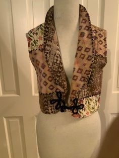 "With the look of a tapestry patchwork, this vest with has a dropped shawl collar and bold frog closure that will take a simple outfit to the next level.  In muted earthtones of brown/beige/peach/pink/greens, it's easy to pair with things already in your closet.  An acrylic blend makes up the tapestry layer, and the back and lining is a light brown cotton.   Bust 34\" Waist 32\" Length 16\"" Bohemian Patchwork Vest For Fall, Fall Bohemian Vest With Patchwork, Fall Bohemian Patchwork Vest, Fitted Beige Bohemian Vest, Bohemian Fitted Beige Vest, Fitted Bohemian Beige Vest, Fitted Multicolor Patchwork Vest, Multicolor Patchwork Fitted Vest, Bohemian Fall Vest Outerwear