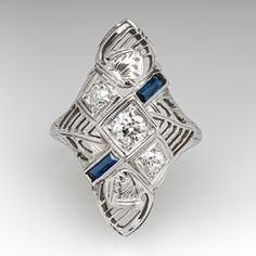 This striking circa 1930s dinner ring features a pierced design with engraved details and bordered with milgrain edging. The ring is accented with three (3), bead set, round transitional brilliant cut diamonds and two (2), bezel set, rectangular French cut synthetic sapphires. The ring measures 23.2mm at the top, rises 5.8mm above the finger, tapering to 1.4mm wide and 0.8mm thick at the base of the shank. This ring is currently a size 3 and can be sized larger. Art Deco Jewelry For Vintage Events, Antique Platinum Three-stone Jewelry, Art Deco Three Stone Platinum Jewelry, Formal Art Deco Three-stone Diamond Ring, Art Deco Jewelry With Center Stone, Vintage Three-stone Platinum Jewelry, Vintage Milgrain Jewelry For Formal Occasions, Art Deco Filigree Ring With Diamond Cut, Vintage Three Stone Platinum Jewelry