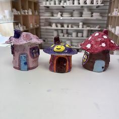 three small clay houses sitting on top of a white counter next to shelves with cups and saucers