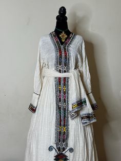 Ethiopian and Eritrean traditional dress featuring intricate embroidery, primarily with geometric patterns in vibrant colors such as red, yellow, green, and black. The embroidery runs vertically down the front of the dress, with additional detailing on the cuffs and lower section of the garment. The fabric appears to be light and flowing, possibly linen or cotton.elegant.beautiful dress. 100%cotton habesha kemis/ netsela and mekeneti Habesha Kemis, Intricate Embroidery, Traditional Dress, Green And Black, Dress 100, Silver Spring, Beautiful Dress, Geometric Patterns, Traditional Dresses