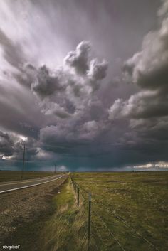 an empty road in the middle of a field with storm clouds above and telephone poles on either side