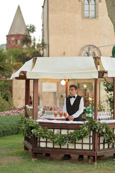 a man standing behind a bar with drinks on it in front of a tall building