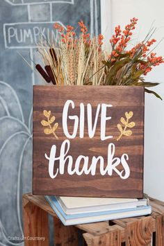 a wooden sign that says give thanks on top of some books and fall leaves in front of a chalkboard