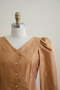 A homemade golden tan jacket, with puffed shoulders, gold faceted buttons, and good tailoring. It is unlined and light enough to be worn as a blouse. ☛ m e a s u r e m e n t s ☚ Bust: 32 Waist: 28 Shoulders: 13.5 Length: 21 ☛ d e t a i l s ☚ Era: 1990s Material: polyester Condition: excellent ☛ v i s i t t h e s h o p ☚ https://etsy.me/2Nd23kg ☛ instagram ┇ poppycockvintage ☛ facebook ┇ poppycockvintage Classic Gold Formal Blouse, Classic Fitted Gold Blouse, Gold Blouse With Buttons For Work, Fitted Gold Top For Workwear, Spring Gold Blouse With Buttons, Fitted Gold Tops For Work, Fitted Gold Blouse For Office, Classic Gold Blouse For Workwear, Gold Formal Blouse With Buttons