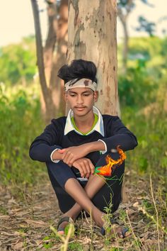 a young man sitting in the grass next to a tree with his hands on his knees