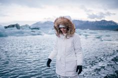 a woman standing in the water wearing a white coat and fur hat with an iceberg behind her