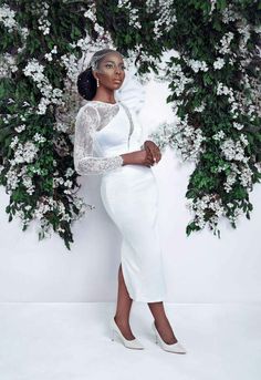 a woman in white is posing for a photo with flowers on the wall behind her