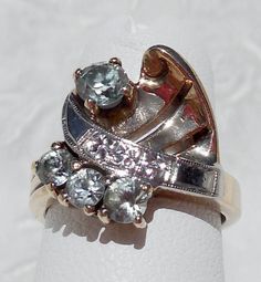 "This great, vintage two-tone ring shows detail and flair typical of the 1940s. The asymmetrical design is composed yellow gold, white gold, and four light blue topaz gemstones. The white gold \"ribbon\" is chased with tiny orange blossoms and decorated with milgrain. It separates the three smaller stones from the one larger stone. The fanned yellow gold section, which shares the side with the larger stone, has two slits and a little curl in the corner. The asymmetry even extends to the shank, w Vintage White Gold Multi-stone Ring, Vintage Multi-stone Diamond Ring Collectible, Vintage Multi-stone Diamond Ring For Collectors, Vintage White Gold Multi-stone Diamond Ring, Vintage Multi-stone Diamond Ring In White Gold, Vintage Multi-stone Sapphire Ring, Vintage Sapphire Ring With Accent Stones, Vintage White Gold Sapphire Ring With Accent Stones, Vintage Three Stone Sapphire Ring