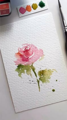 a rose painted with watercolors on white paper next to some paintbrushes