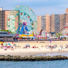 many people are on the beach in front of an amusement park with ferris wheel and rides