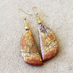 Handmade carved Picasso Jasper earrings suspended from a raw brass ear hook.  Stone: Carved Piscasso Jasper Colour: Browns, Oranges & Yellows  Metal: Hammered raw brass, lead and nickel free Earrings Drop Length: 7.5cm Earrings Width: 1.8cm I only make one of each pair of earrings, so each piece is as unique as you.  Designs can be customised specifically to meet your needs with your favourite semi-precious stone. Please contact me about a custom design. Handmade Jasper Gold Jewelry, Handmade Gold Jasper Jewelry, Gold Nature-inspired Earrings With Natural Stones, Nature-inspired Gold Earrings With Natural Stones, Gold Natural Stones Earrings Nature-inspired, Gold Jasper Jewelry For Jewelry Making, Earthy Brass Dangle Earrings, Nature-inspired Gold Wire Wrapped Earrings, Artisan Earrings With Natural Stones And Brass