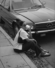 a woman sitting on the curb drinking from a bottle next to a car with its hood up