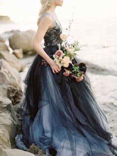 a woman in a black and blue dress is standing on rocks near the water holding a bouquet