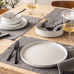 a wooden table topped with white plates and silverware next to two glasses of wine