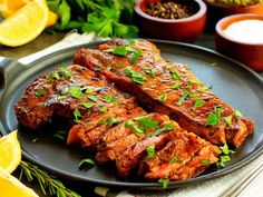 two pieces of meat on a black plate with lemons and seasoning in the background