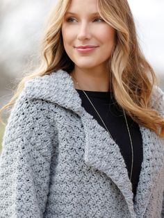 a woman wearing a gray crochet coat and black top smiles at the camera