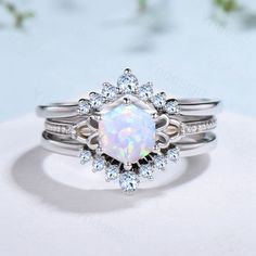 a white opal and diamond ring set on top of a white surface with flowers in the background