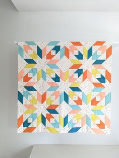 a colorful quilt hanging on the wall next to a white shelf with an orange, blue, and yellow geometric design