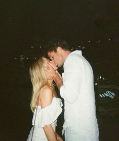 a man and woman kissing each other in front of the ocean at night time with city lights behind them