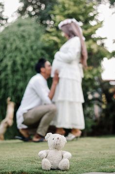 a man kneeling down next to a white teddy bear in the grass with a woman standing behind him