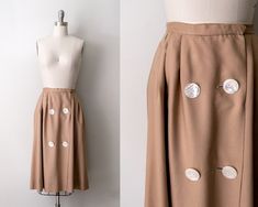 "1950's tan rayon skirt. Double set of large mother-of-pearl white buttons down the front. Metal eye and hook clasps at front. Pleats on hips. Mid-length hem with pink lining. Dimensions: Waist 25\" Hips open Length 27\" Fits Like: Small Era: 1950's Label: Suburbia USA Condition: Great vintage. Cleaned and ready to wear. All clothing measurements are taken flat, lying down and then doubled (when needed). Any stretching or other specifications, if any, will be noted next to individual measurement Vintage Brown Skirt With Buttons, Vintage Full Skirt Bottoms With Buttons, Vintage Full Skirt With Buttons, Vintage Brown Bottoms With Buttons, Vintage Fall Skirt With Buttons, Vintage Full Skirt Bottoms In Beige, Vintage Beige Full Skirt Bottoms, Vintage Fitted Skirt With Buttons, Fitted Vintage Skirt With Buttons