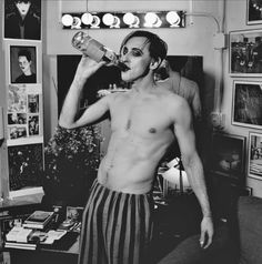 a shirtless man drinking from a bottle while standing in a room with pictures on the wall