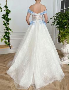 a woman in a white wedding dress with blue flowers on the waist and shoulder, standing next to a potted plant