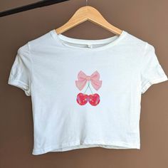 Introducing the Coquette Cherry Ribbon Bow Women's Crop Tee, a must-have addition to your wardrobe for those who appreciate softcore balletcore era fashion and trendy vintage cute aesthetic. This adorable t-shirt combines the elegance coquette with the charm of a holographic set of cherries with a ribbon bow. Made from high-quality materials, this crop tee offers both comfort and style. Embrace your inner princesscore art enthusiast and make a fashion statement with this versatile piece. Whether you pair it with high-waisted jeans for a casual yet chic look or with a skirt for a more polished ensemble, this crop tee is sure to turn heads. The soft fabric feels gentle against your skin, while the bow adds a touch of whimsy to your outfit. The Coquette Cherry Ribbon Bow Women's Crop Tee is p White Cotton Aesthetic T-shirt, Cute White Cotton Cropped T-shirt, White Y2k Tops With Cherry Print, Aesthetic Graphic Print Tops For Summer, Aesthetic Summer Tops With Letter Print, Aesthetic Graphic Print Tops For Spring, Cute Summer Crop Top With Funny Print, Aesthetic Short Sleeve Tops With Letter Print, Aesthetic Letter Print Summer Tops