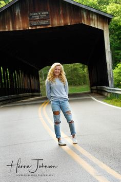 a woman standing on the side of a road in front of a wooden covered bridge