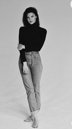 a woman standing in front of a white background wearing jeans and a turtle neck sweater