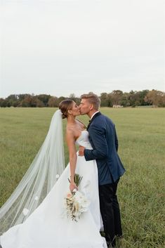 a bride and groom kissing in the middle of a field with their veil blowing in the wind